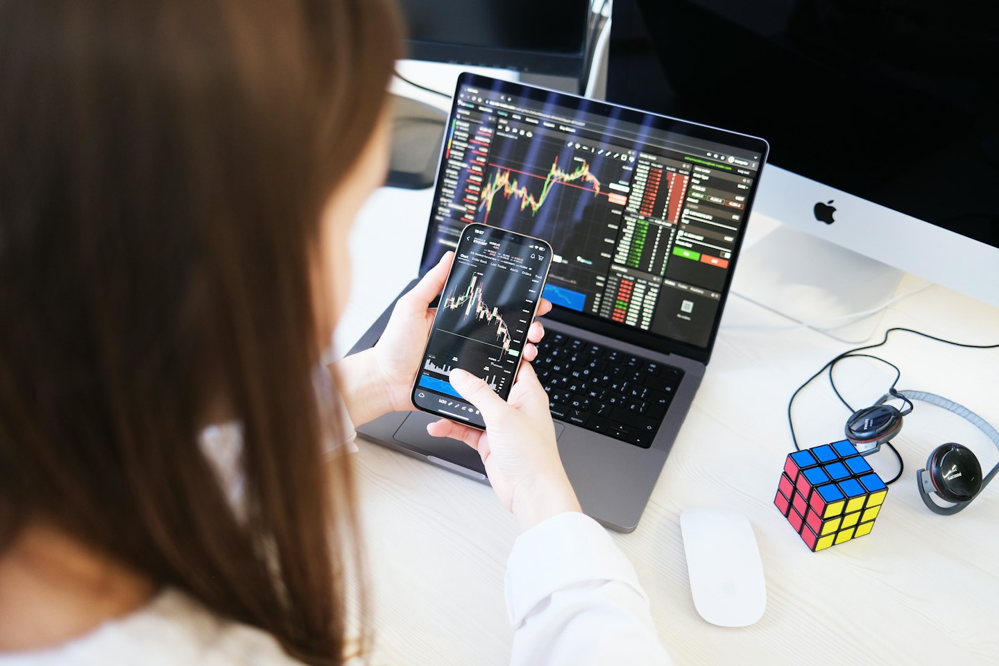 Person trades stocks on phone and laptop in office.